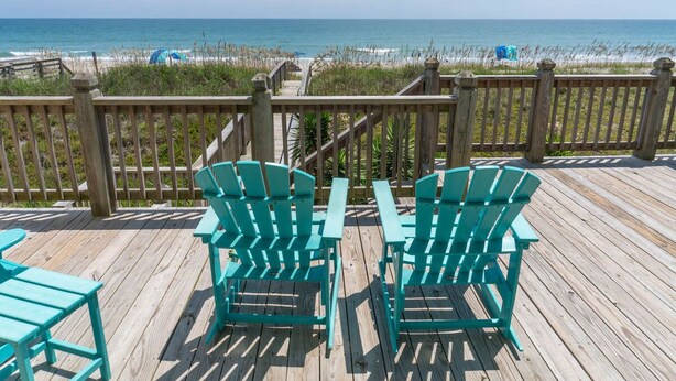 A wooden deck with turquoise Adirondack chairs and a table, overlooking a beach with a wooden railing and distant beach chairs. - Dune Alright, Oceanfront House in Emerald Isle, NC! (Emerald Isle)