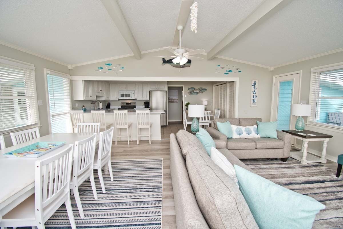 A spacious living area with a sofa, dining table, and chairs, a kitchen in the background, and a ceiling fan.