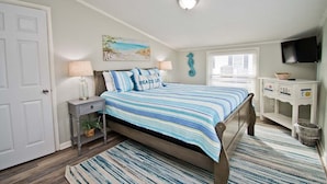 A bedroom with a bed, bedside tables, a lamp, a painting, a TV, and a window with blinds. - Dune It Whale, Oceanfront House In Emerald Isle, NC! (Emerald Isle)