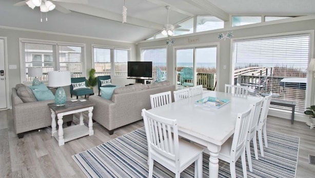A spacious living room with a large white dining table, a sofa, and a TV. - Dune It Whale, Oceanfront House In Emerald Isle, NC! (Emerald Isle)