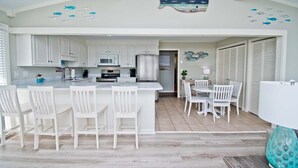 A modern kitchen with white cabinetry, a central island with bar stools, and a dining area with white chairs. - Dune It Whale, Oceanfront House In Emerald Isle, NC! (Emerald Isle)
