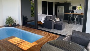 Un área de estar moderna con una piscina, un sofá, una mesa de comedor y una cocina visible a través de un vidrio.