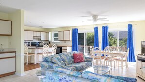 A spacious living area with a sofa, dining table, and chairs, a kitchen with cabinets and appliances, and a balcony with blue curtains. - Dune Magic, Almost Oceanfront House in Emerald Isle, NC! (Emerald Isle)