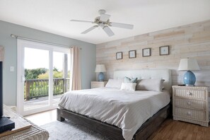 Une chambre à coucher avec un lit, deux tables de chevet, un ventilateur de plafond, un mur en bois et un balcon avec vue.