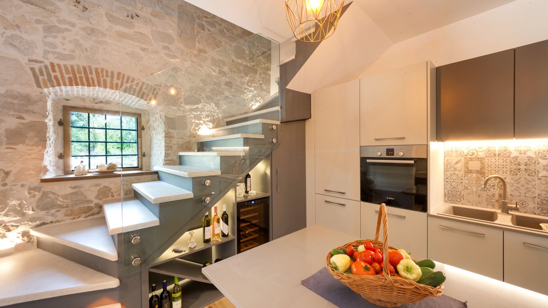 A modern kitchen with a stone wall, a built-in staircase, and a large window.