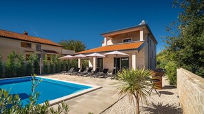 A poolside area with lounge chairs and umbrellas, a swimming pool, and a building with a tiled roof.