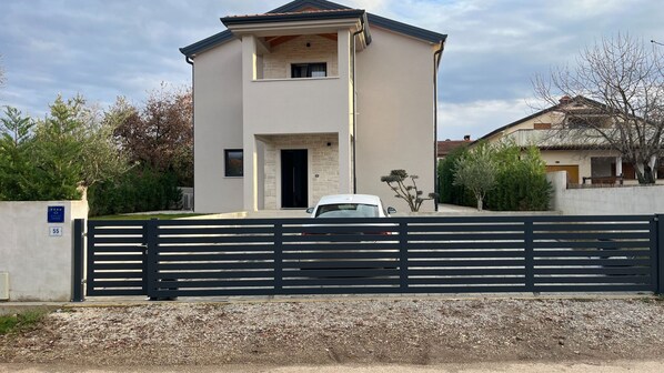 A modern two-story house with a gated driveway, a visible garage door, and a small balcony.