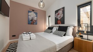 Ein modernes Schlafzimmer mit einem großen Bett, zwei an der Wand befestigten Bildern, einem Nachttisch mit Lampe und einem Fenster mit Blick.