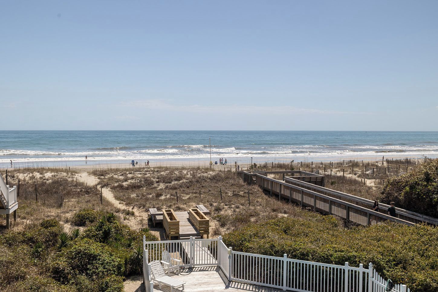 A beach view with a wooden boardwalk, beach chairs, and a fence.