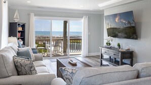A living room with a sofa, a coffee table, a bookshelf, a TV, and a balcony with a view of the ocean. - Footprints East, Oceanfront Duplex in Emerald Isle, NC! (Emerald Isle)
