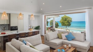 A modern living room with a large window overlooking a beach, a kitchen with white cabinets, and a dining area with wooden furniture.