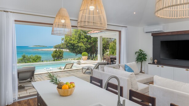 A modern kitchen with a view of the sea, a dining area with a white table, and a cozy seating area with a TV.