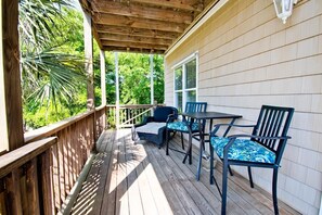 A covered outdoor patio with a wooden deck, a table with chairs, and a view of greenery. - Grace Appearing, Oceanside House in Emerald Isle, NC! (Emerald Isle)