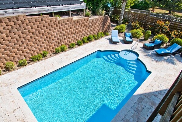 A rectangular swimming pool with a blue mosaic pattern, surrounded by a brick wall, a wooden deck, and lounge chairs.