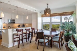 A modern kitchen with white cabinetry, a central island with bar stools, a dining table with chairs, and a large window with a view. - Grande Villas A-3, Oceanfront Condo in Indian Beach, NC! (Indian Beach)