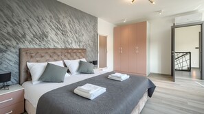 Une chambre moderne avec une tête de lit capitonnée, un grand lit agrémenté de nombreux coussins, une couvre-lit grise à motifs et une armoire aux portes coulissantes.