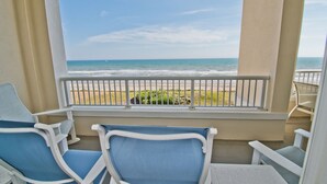 Un balcon avec deux chaises bleues et une petite table, d’où l’on a une vue dégagée sur une plage, l’océan et la côte lointaine.
