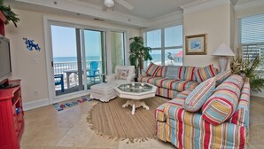 A living room with a large window, a ceiling fan, a striped sofa, a glass coffee table, and a red cabinet. - Grande Villas M-3, Oceanfront Condo in Indian Beach, NC! (Indian Beach)