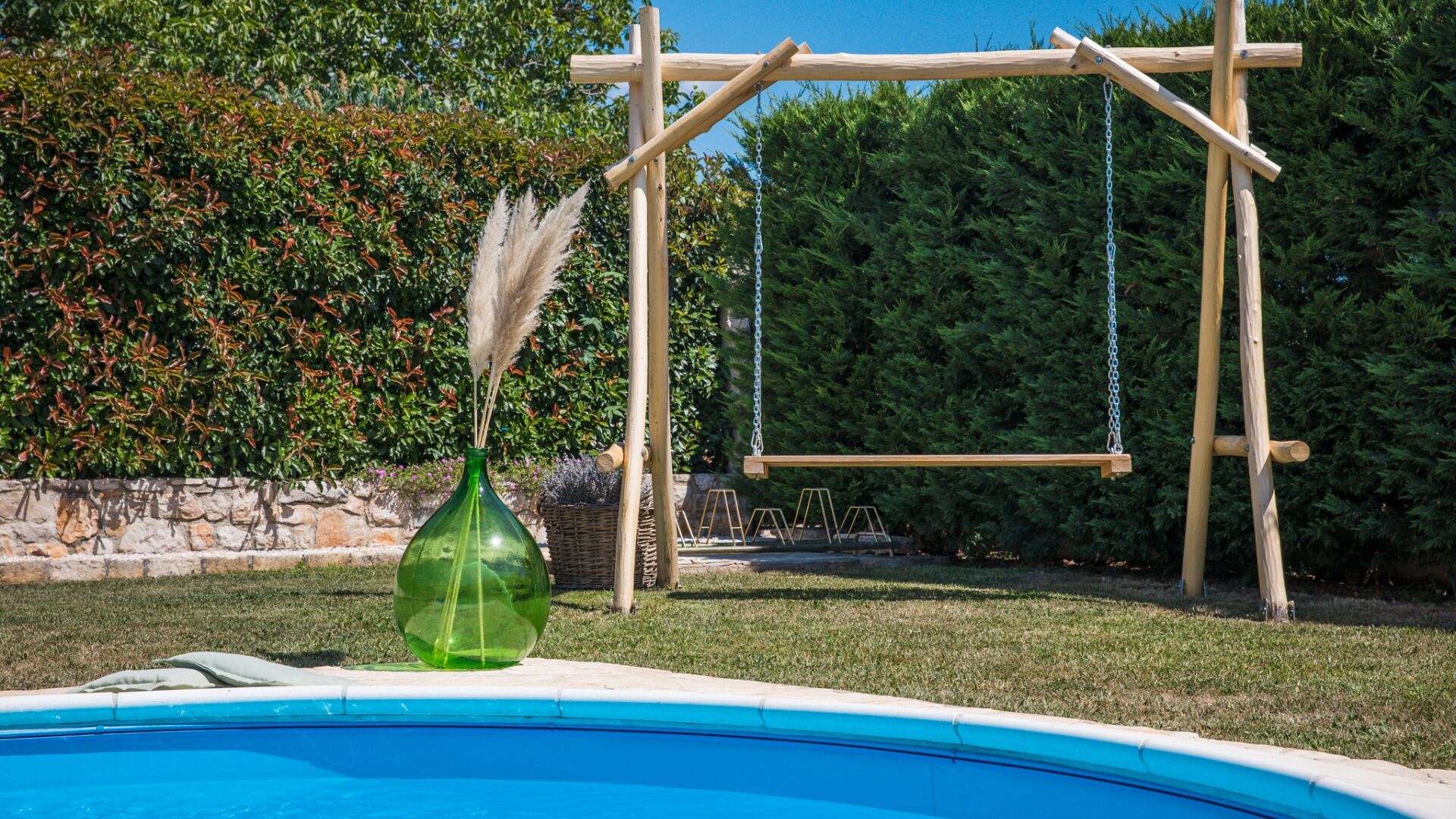 A pool with a wooden swing set and a green vase with pampas grass.