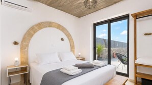 Quarto com cama grande, um banco, uma mesinha e vista para o exterior.