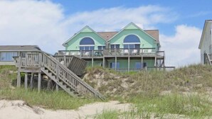 Een strandhuis met een houten trap naar een terras, een groot, dubbel hoog gebouw met een groene gevel en een zandstrand op de voorgrond.