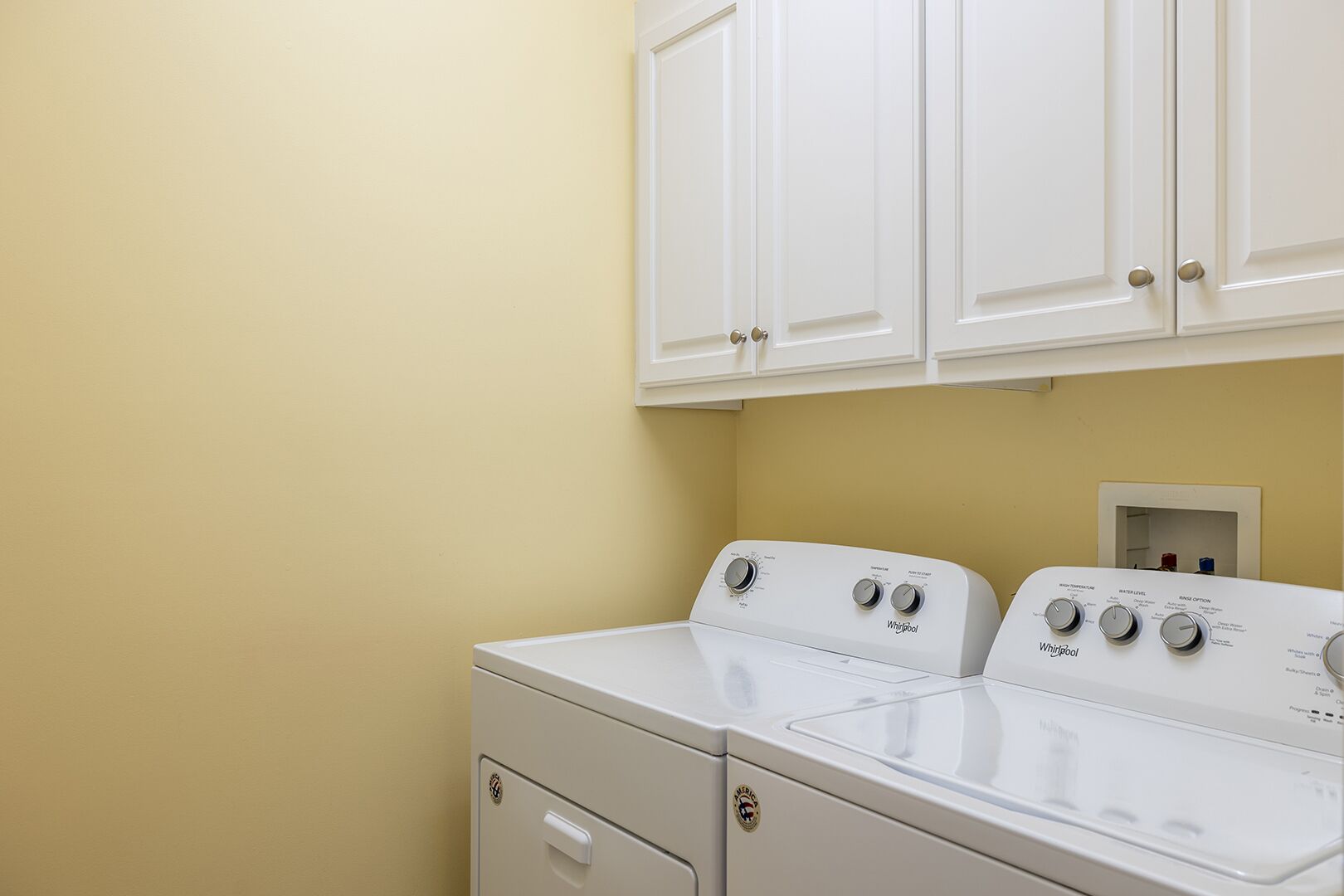 A laundry room with two white Whirlpool washers and dryers, white cabinets above, and a beige wall.