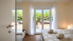 A hotel room with a bed, a nightstand, a window with curtains, and a view of parked cars outside.