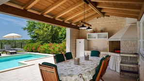 Un espacio al aire libre cubierto con piscina, mesa de comedor, sillas y zona de cocina con pared de piedra y armarios.