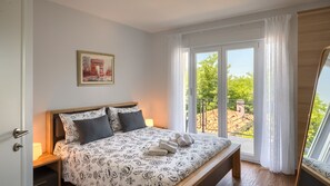 A bedroom with a bed, a bench, a nightstand, a lamp, a window with curtains, and a framed picture on the wall.