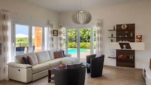 Una sala de estar con sofá, sillón y mesa de centro, además de una estantería y vistas a una piscina.