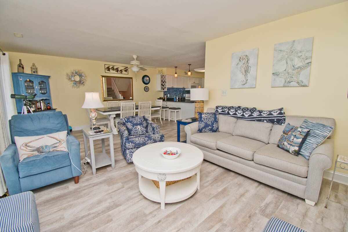 A living room with a blue armchair, a beige sofa, a white coffee table, and a dining area with a white table and chairs.
