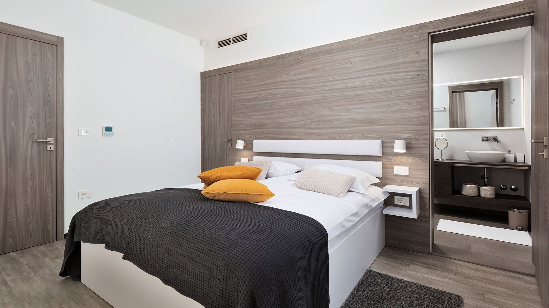 A modern bedroom with a bed, bedside tables, a nightstand, a mirror, and a bathroom with a sink and toilet.