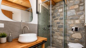 Un baño con tocador de madera, un lavamanos blanco, cabina de ducha de cristal y pared de piedra.