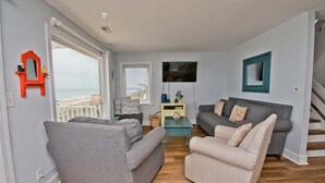 A living room with two grey sofas, a small coffee table, and a TV. There is a window with blinds, a red mirror, and a staircase. - Tides Out West, Oceanfront Duplex in Emerald Isle, NC! (Emerald Isle)