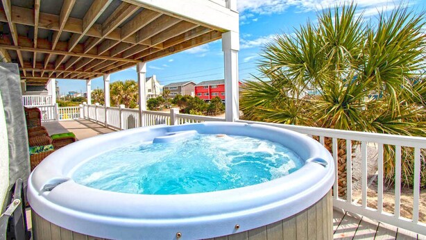 Jacuzzi sobre un pont de fusta amb barana blanca, envoltat de palmeres i un cel blau clar.