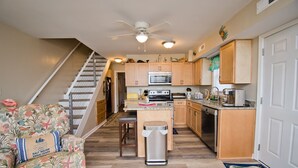 A compact kitchen with wooden cabinets, a microwave, and a stove. There's a small island with a sink and a trash can. A chair with a floral pattern is visible, and a staircase leads to another level. - Tranquillis East, Almost Oceanfront Duplex in Emerald Isle, NC! (Emerald Isle)