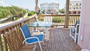 A wooden deck with a glass table and chairs, overlooking a residential area with houses and a beach. - Tranquillis East, Almost Oceanfront Duplex in Emerald Isle, NC! (Emerald Isle)