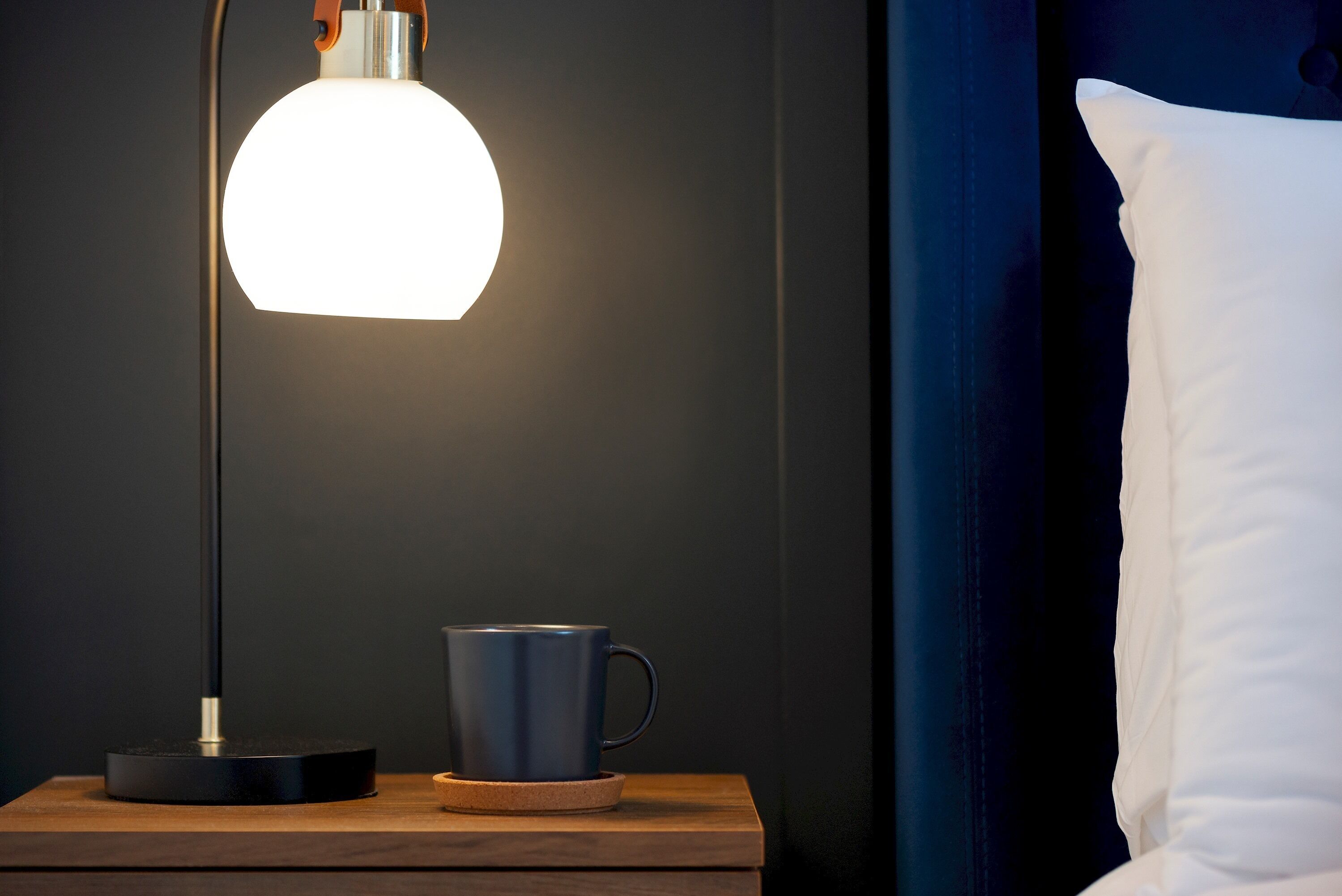 Una mesita de noche con una lámpara, una taza y un almohadón sobre una pared oscura.