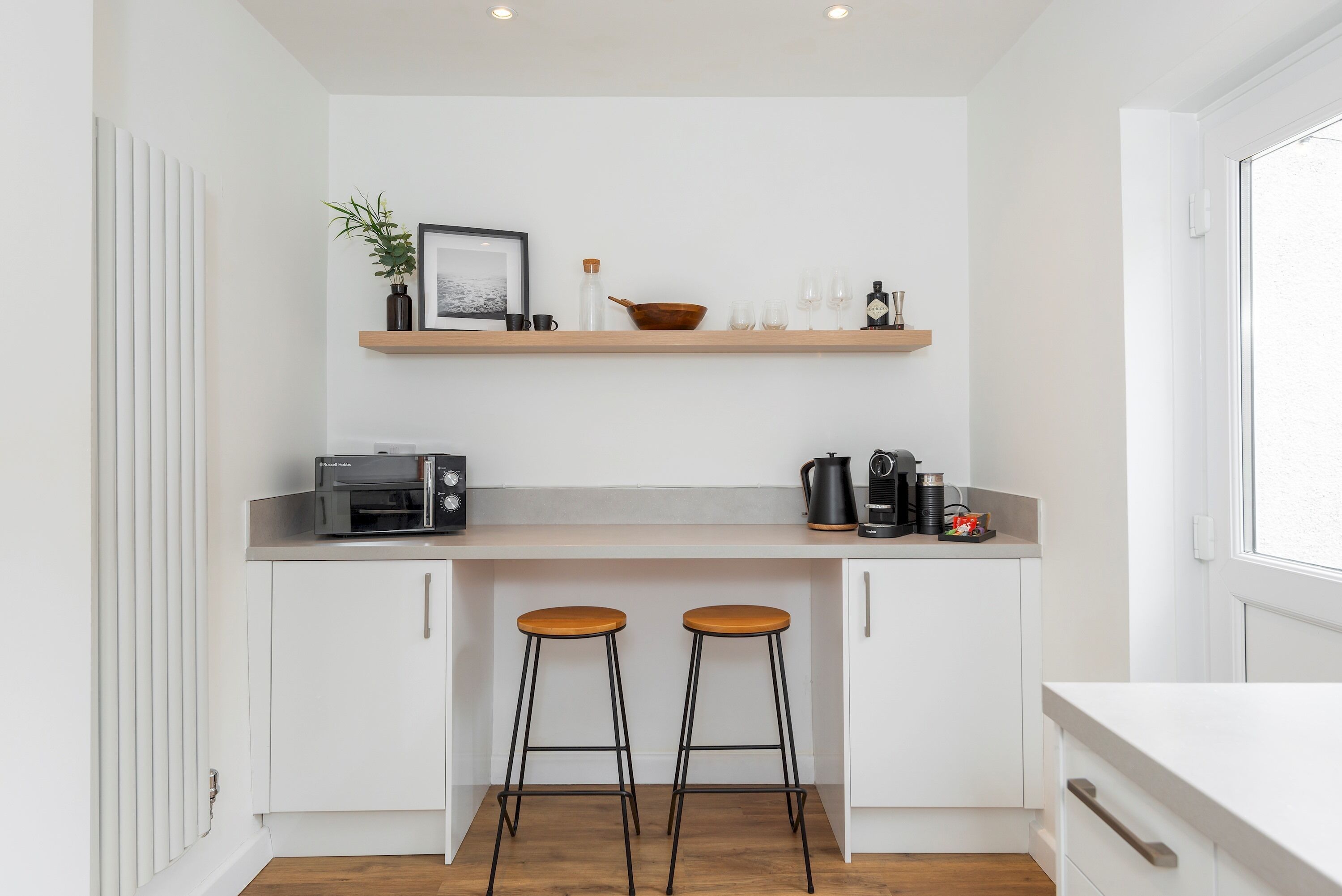 Cocina pequeña con armarios blancos, encimera de madera, cafetera y dos taburetes.