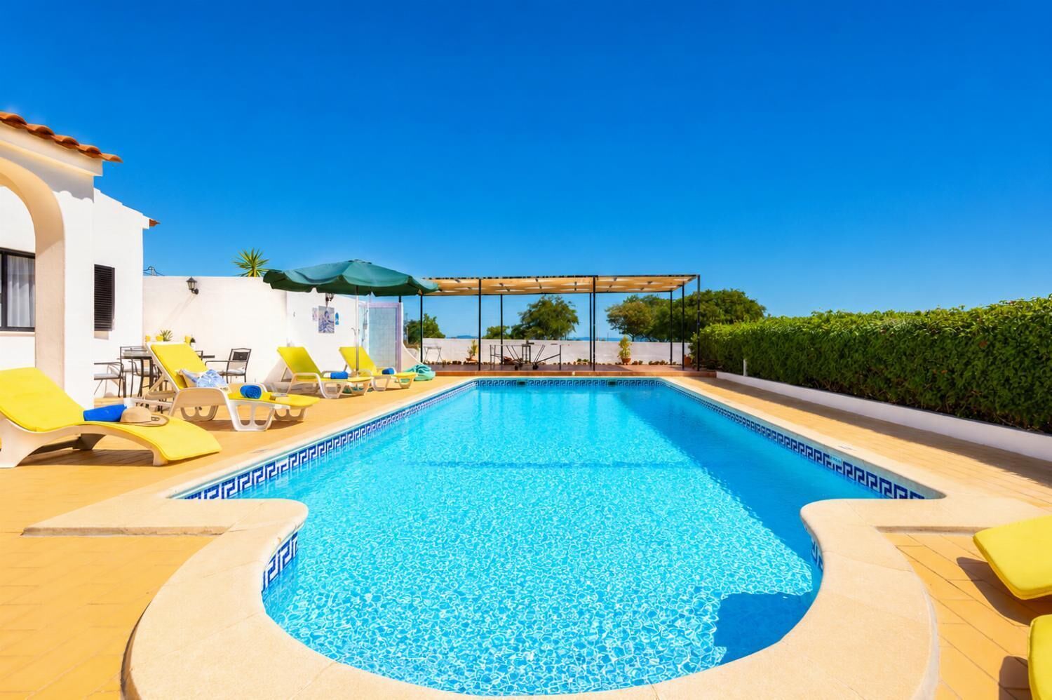 Ein Swimmingpool mit gelben Liegestühlen, einer Pergola und einem Gebäude mit weißem Putz und einem Ziegeldach.