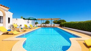 A swimming pool with yellow lounge chairs, a pergola, and a white building with a tiled roof.