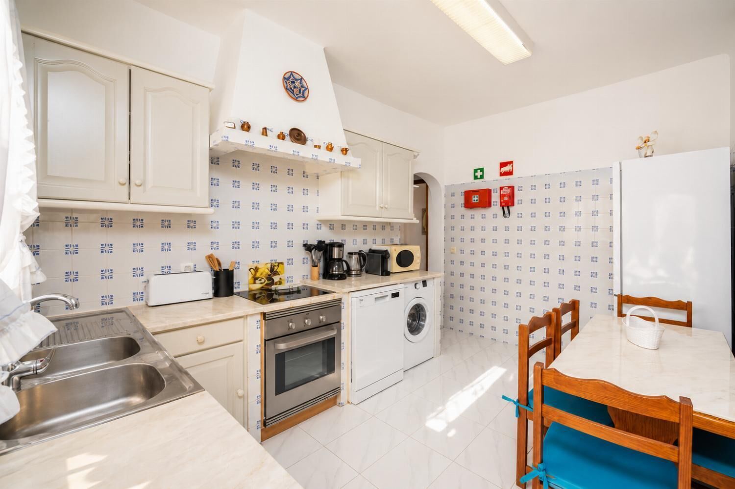A compact kitchen with a sink, stove, oven, and washing machine. There is a dining table with chairs and a refrigerator.