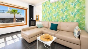 Een moderne woonkamer met een hoekbank, een tafeltje met een hoed, een groot raam met uitzicht en een wand met een tropisch bladpatroon.