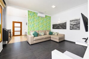Un salon moderne avec un canapé d’angle beige, un grand mur orné d’un motif de feuilles tropicales et des tableaux représentant des paysages en noir et blanc.