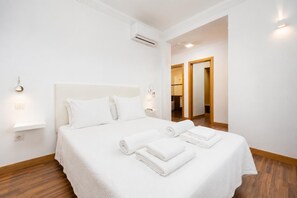 A hotel room with a large bed, white linens, a nightstand, wall-mounted lights, and a wooden floor.