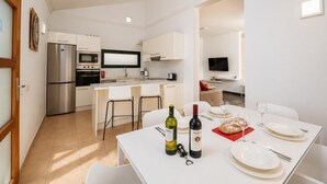 Cozinha moderna com forno embutido, micro-ondas e frigorífico. Dispõe de zona de refeições com mesa posta para dois, incluindo garrafas de vinho e pão.