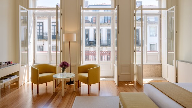 Una habitación con una cama, dos sillones amarillos, una mesita con un jarrón y ventanas amplias que dan a edificios.