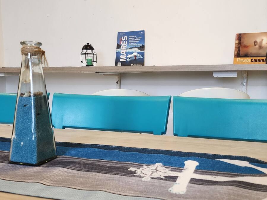 Un salón de conferencias con una mesa larga, sillas azules, una botella de vidrio, un farol y una estantería con un mapa y un control remoto.