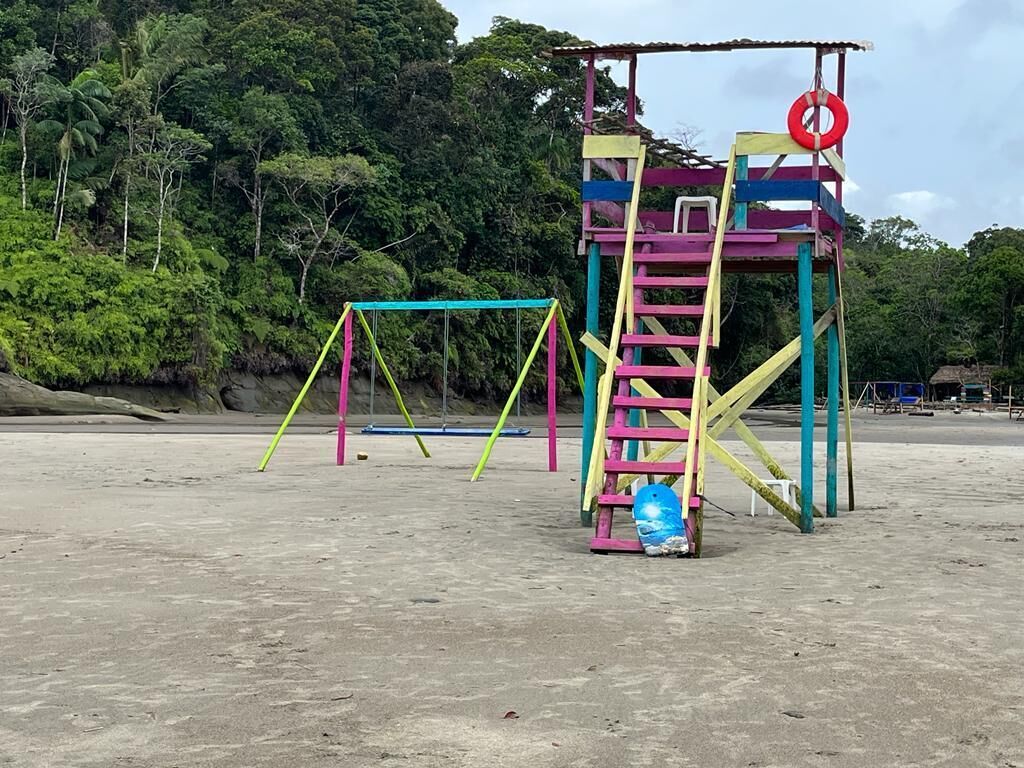 A playground with colorful structures and swings on a sandy beach with dense greenery in the background.