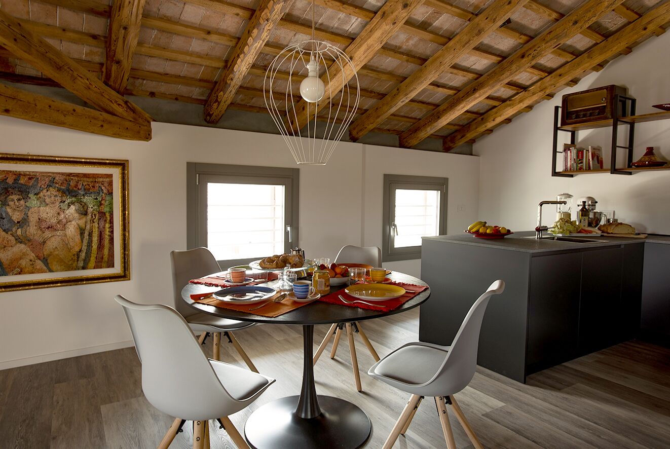 A dining area with a round table, white chairs, and a hanging light fixture. There is a painting on the wall and a kitchen area with a counter and appliances.